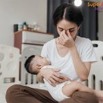 Adult sitting on nursery floor holding baby, hand covering face in moment of stress or fatigue