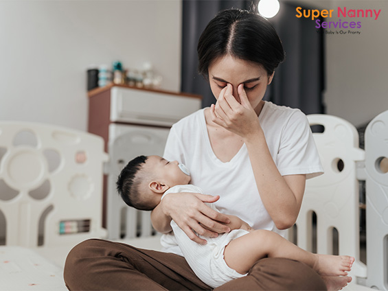 Adult sitting on nursery floor holding baby, hand covering face in moment of stress or fatigue
