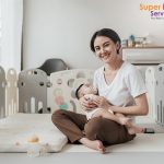 Asian mother sitting on a mat holding a baby in a play area.