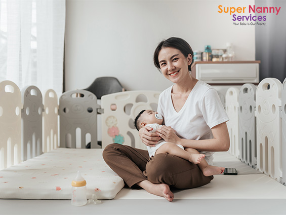 Asian mother sitting on a mat holding a baby in a play area.