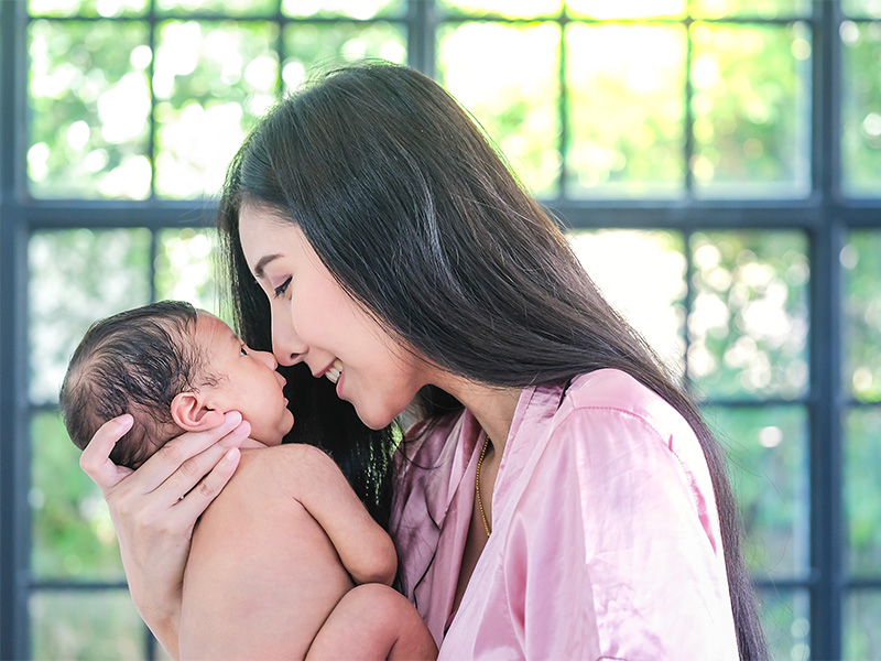 Adult holding newborn close with faces touching, framed by window light and greenery in background.